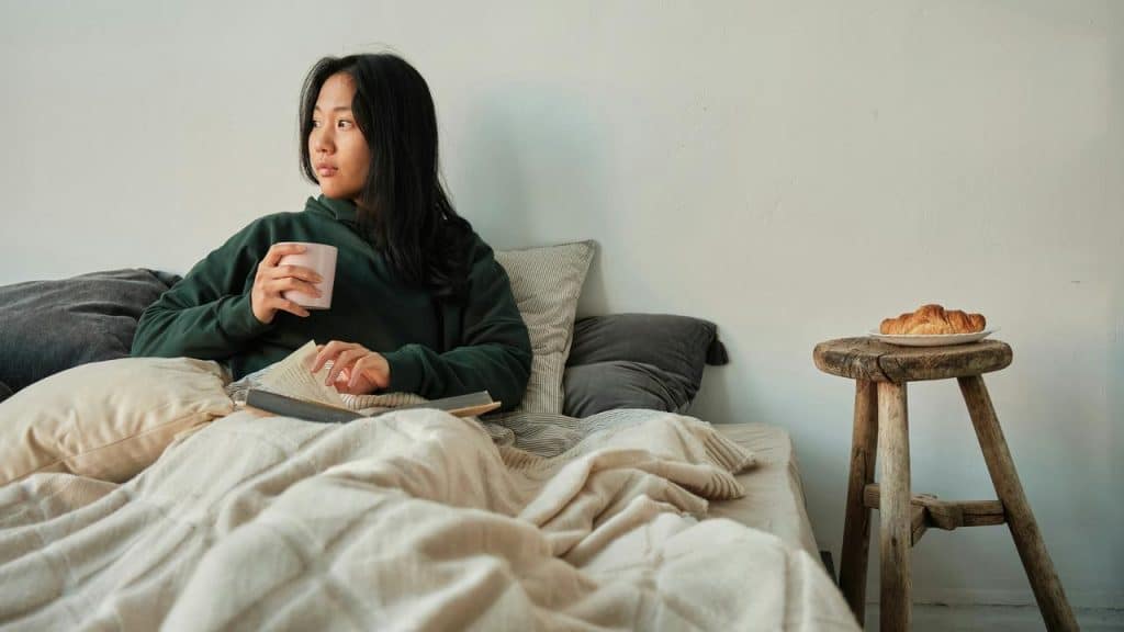 A person sitting up in bed holding a mug and reading beside a small stool with a pastry.