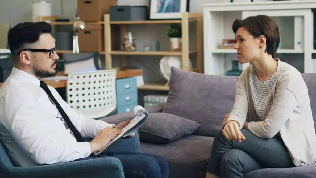 A man with a clipboard listens to a woman sitting on a gray couch.