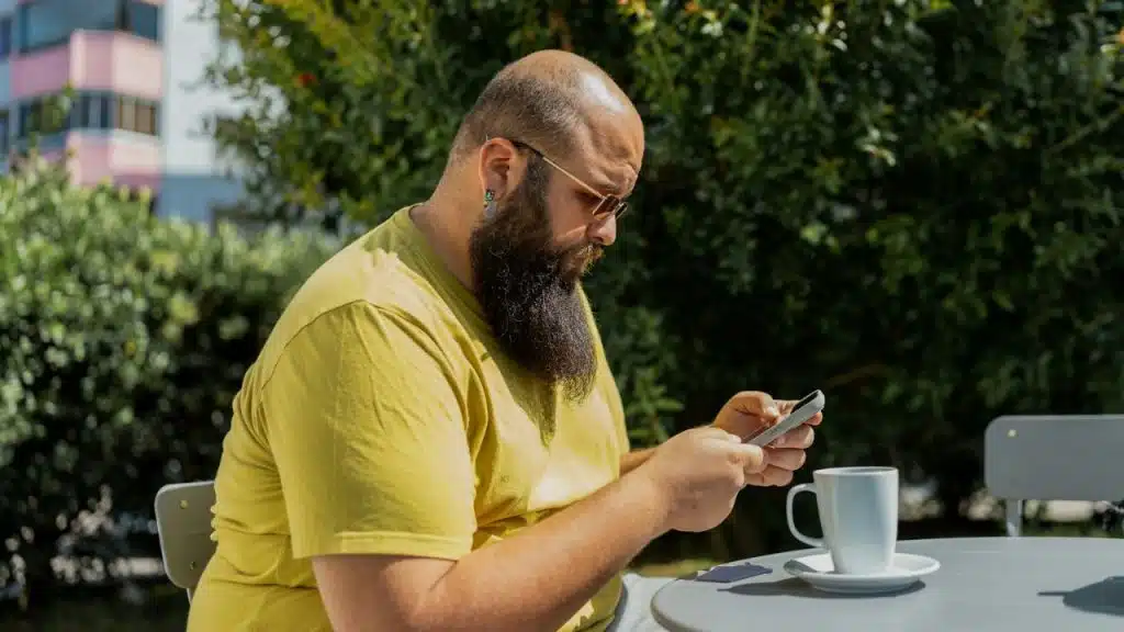 A man with a large beard and sunglasses looks at a smartphone at an outdoor table.