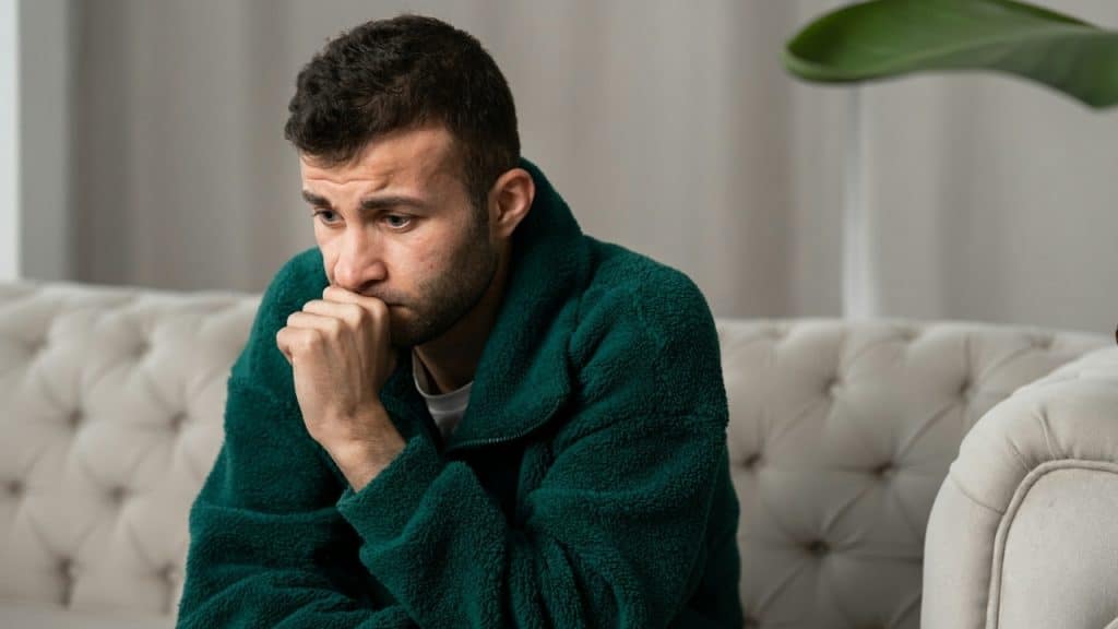 A man in a green fleece jacket sits on a sofa with his chin on his hand.