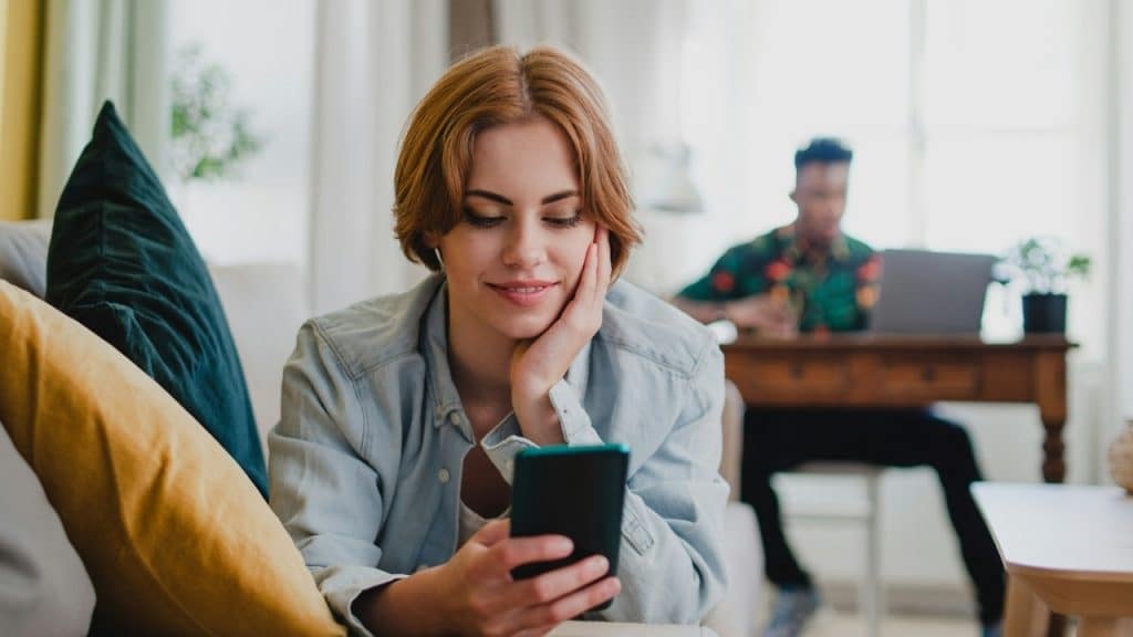 A woman smiles while looking at her phone as a man works in the background.
