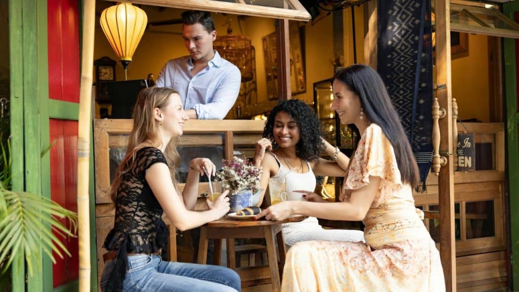Three women sit at a small outdoor table while a man stands behind a counter.
