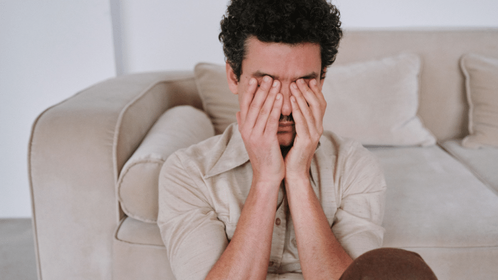 A man with curly hair sits on the floor, covering his face with both hands.