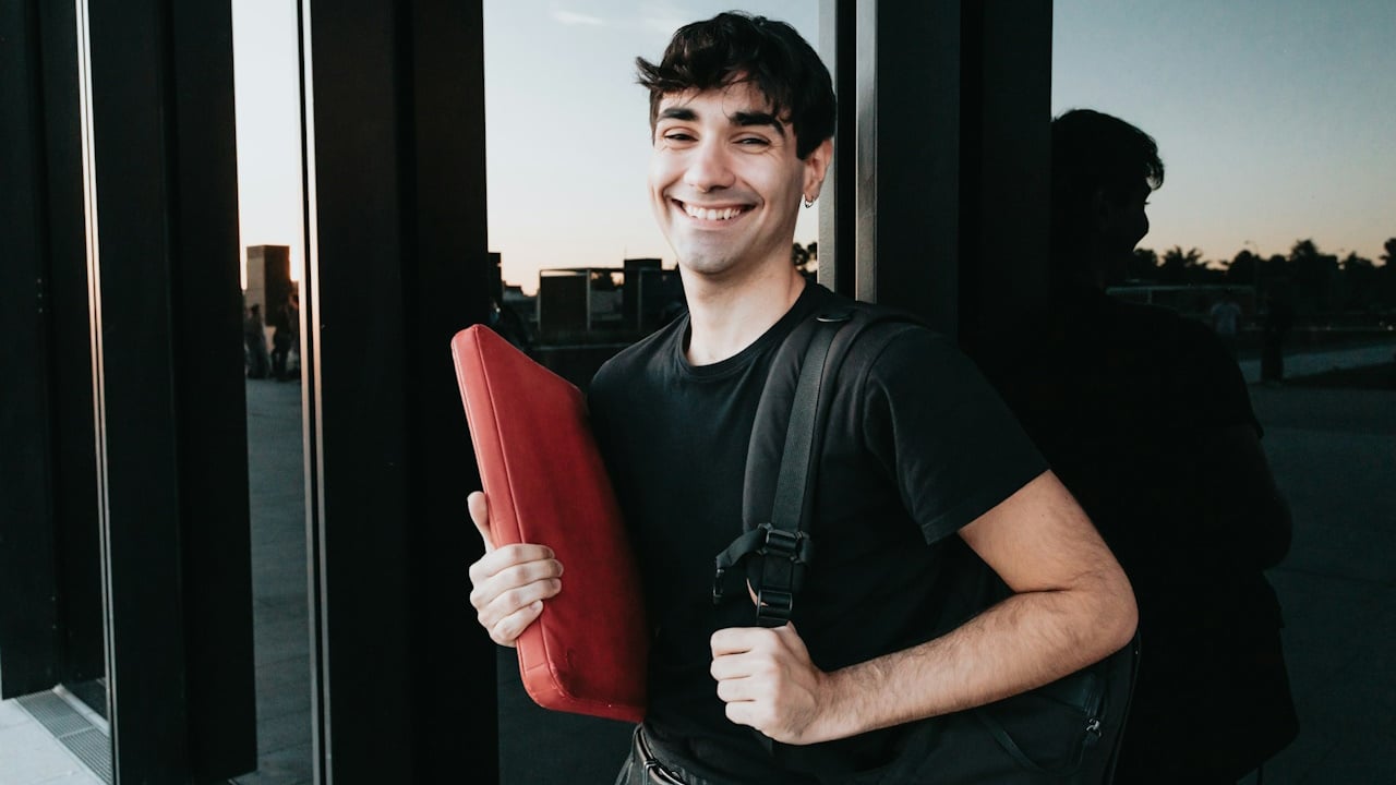 A man holding a laptop and a backpack is giving a smile to the camera.