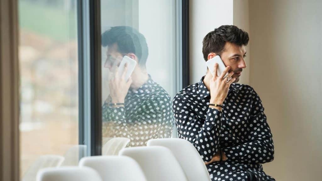A man in a polka-dot shirt talks on a phone by a window, reflected in the glass.