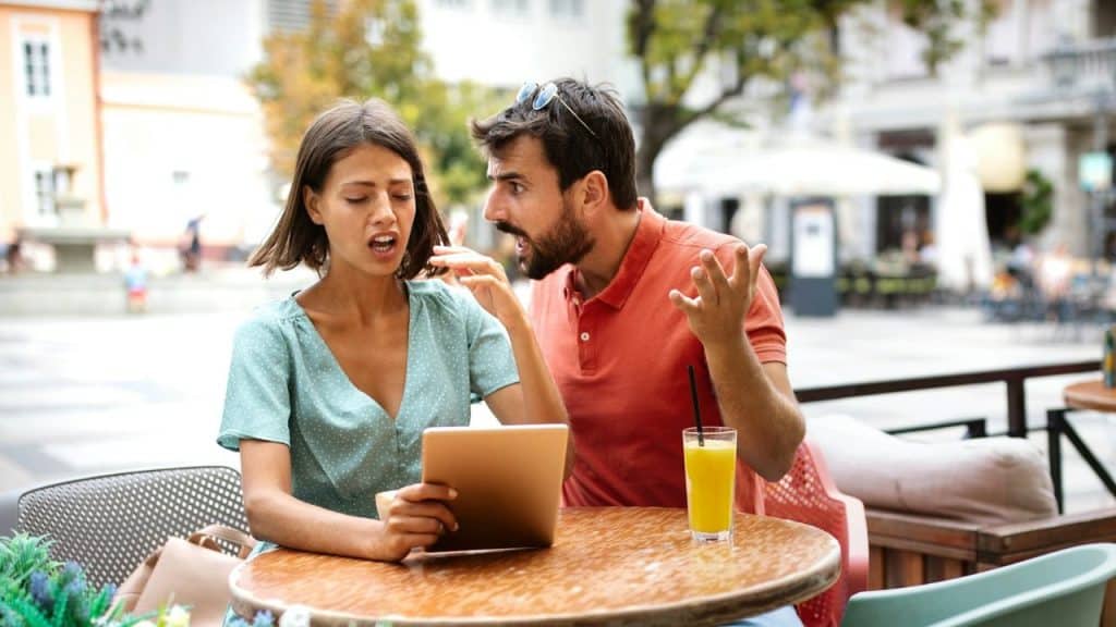 A man gestures broadly at a woman holding a tablet while sitting at outdoor table.