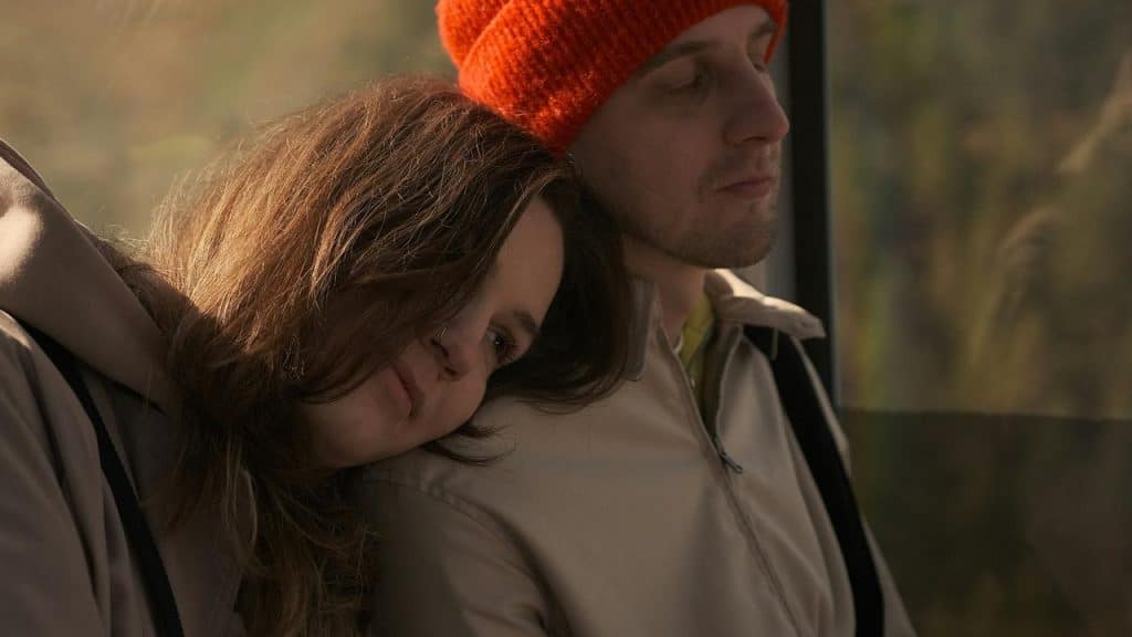 A woman resting her head on a man's shoulder while he wears an orange beanie outdoors.