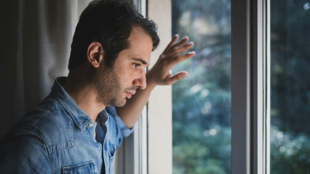 A man in a denim shirt leans against a window and looks outside with a somber expression.