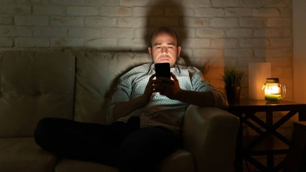 A man sits on a sofa in a dark room looking at a glowing smartphone screen.