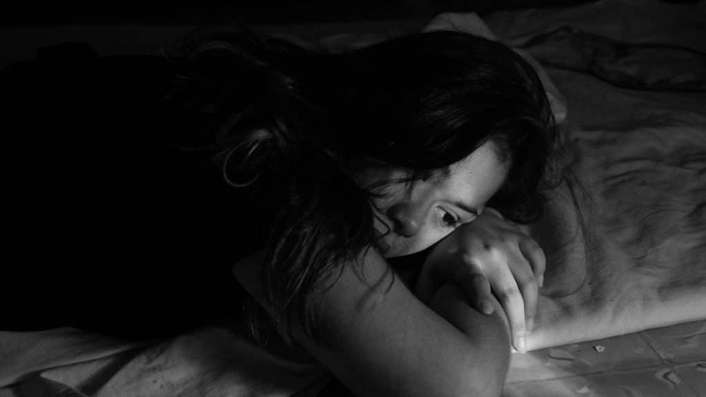 A black and white image of a woman lying on a bed with her head resting on her arms, looking sad or troubled.