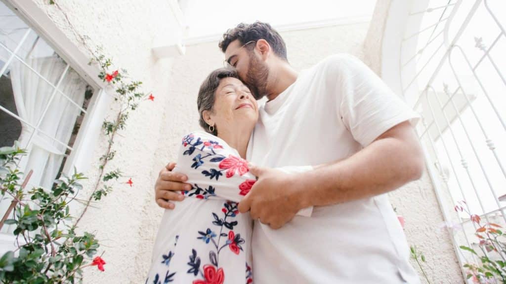 A man with a beard hugs an older woman and kisses the top of her head.