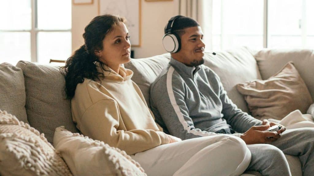 A woman sits on a couch next to a man wearing headphones and holding a controller.