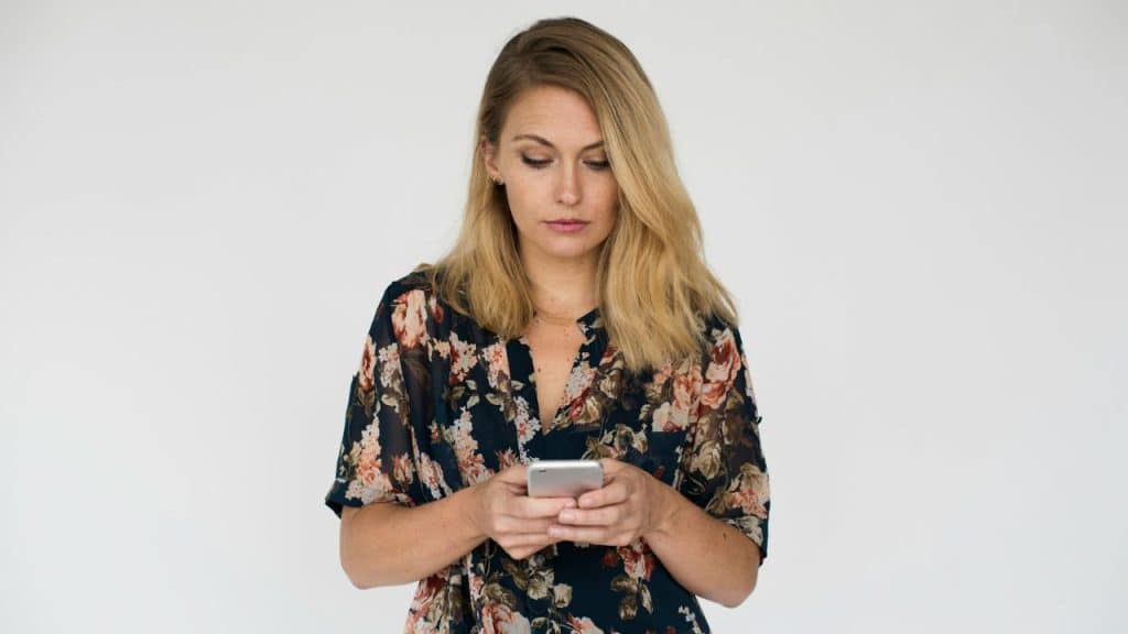 A woman in a dark floral shirt looks down at a smartphone against a white background.