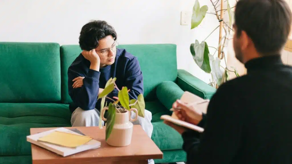 A person on a green sofa rests their head on their hand, looking away from another person.