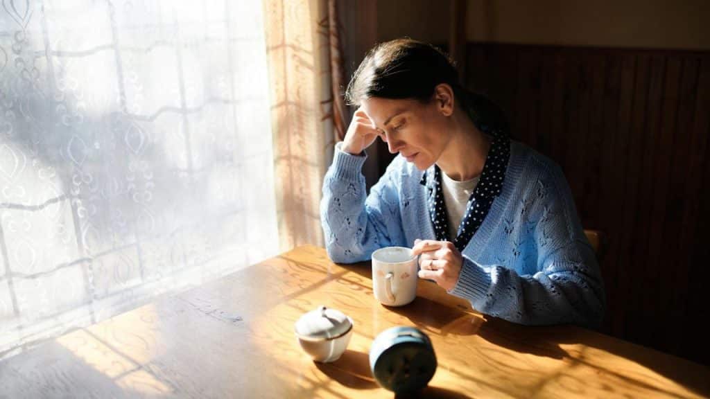A pensive woman sits alone at a wooden table in sunlight, holding a mug with her hand resting on her head.
