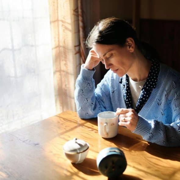 A pensive woman sits alone at a wooden table in sunlight, holding a mug with her hand resting on her head.