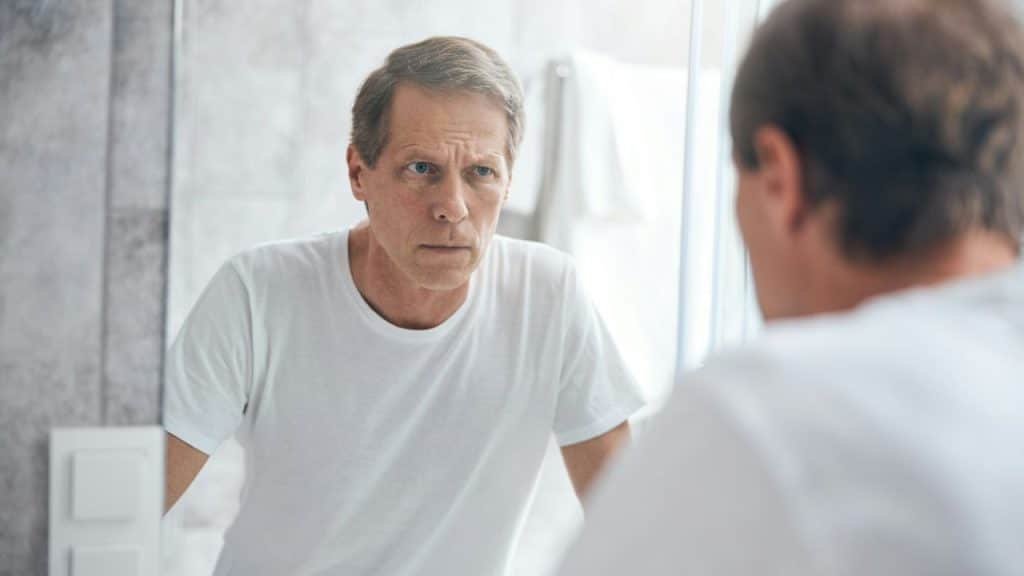 A man in a white t-shirt looks at his own reflection in a bathroom mirror.