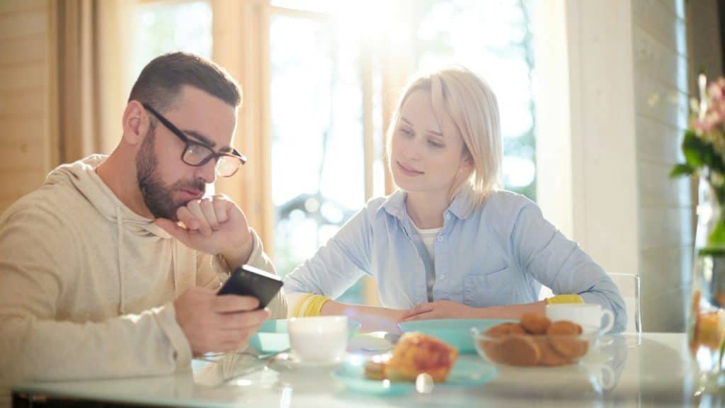 A man and woman sit at a table while the man looks at his smartphone.