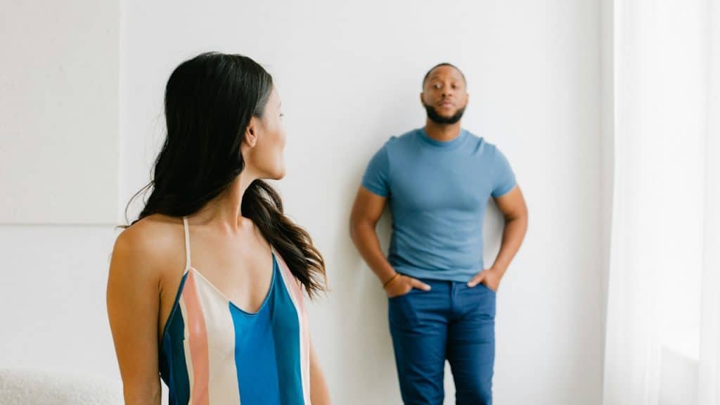 A woman staring back at a bearded man leaning against a wall with his hands in his pockets.