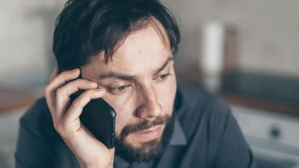 A man with a beard holds a smartphone to his ear while looking downward thoughtfully.