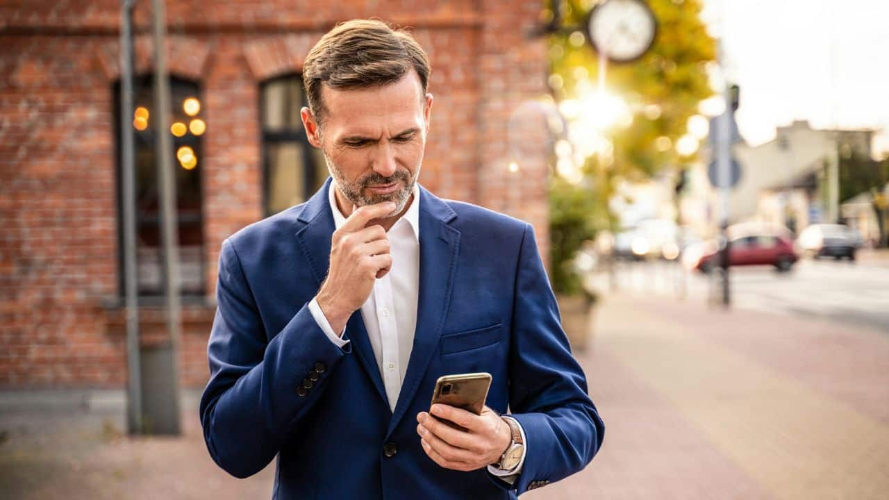 A man in a blue suit stands outside looking at his phone with a concerned expression.