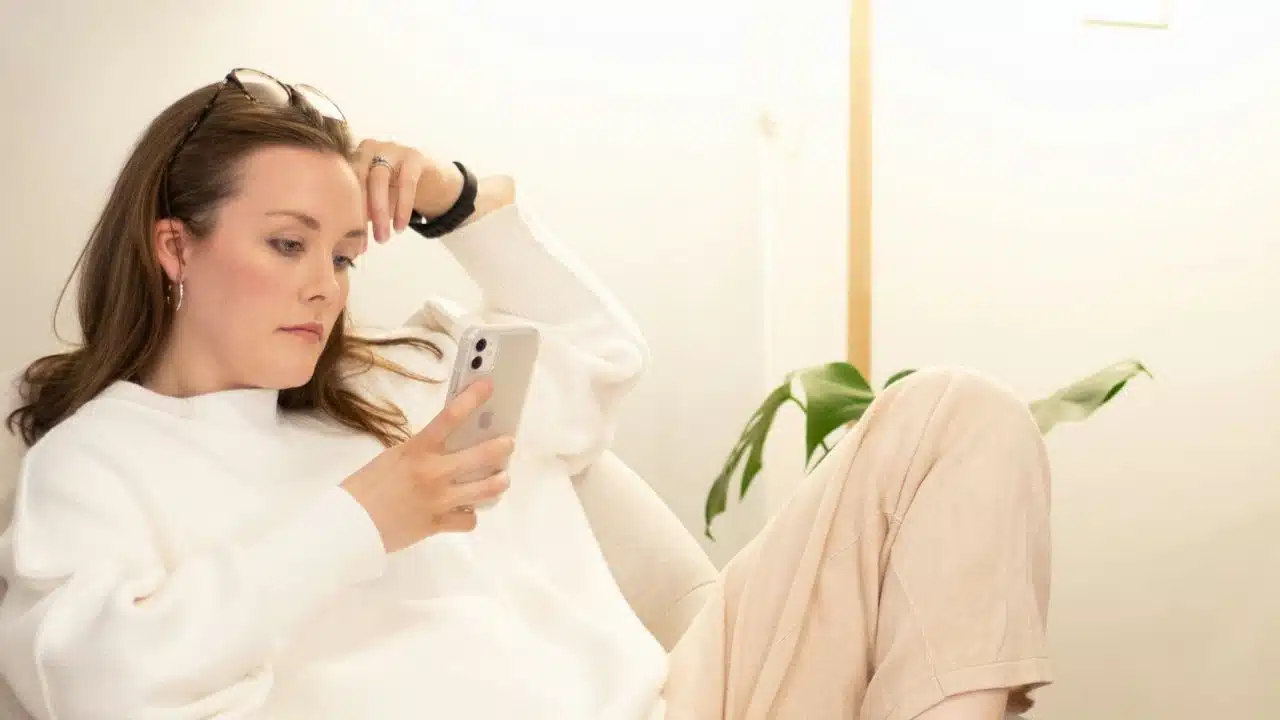 A woman in a white sweater reclines while holding a smartphone and looking at the screen.