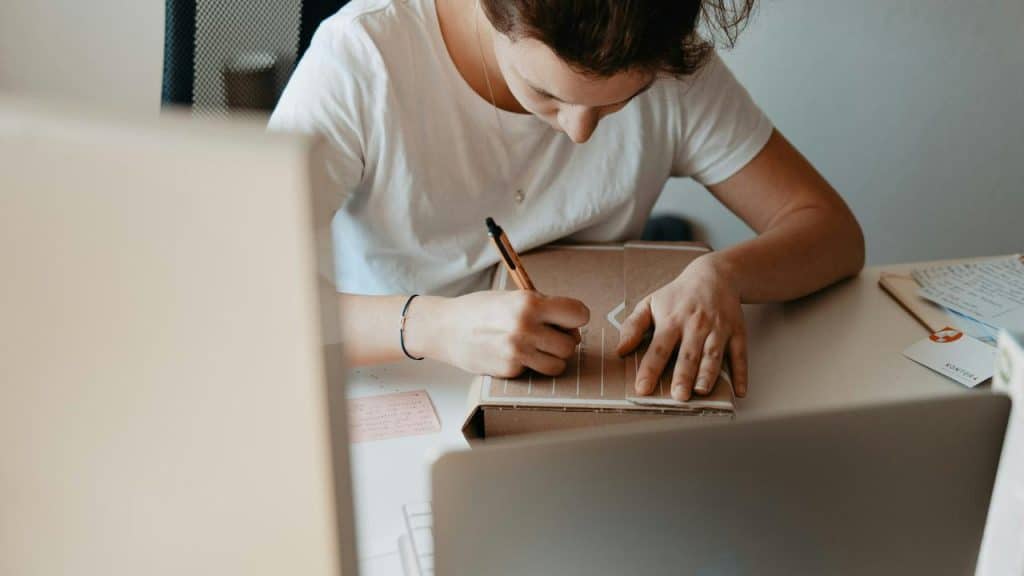 A person writing on a cardboard box at a desk with a laptop.