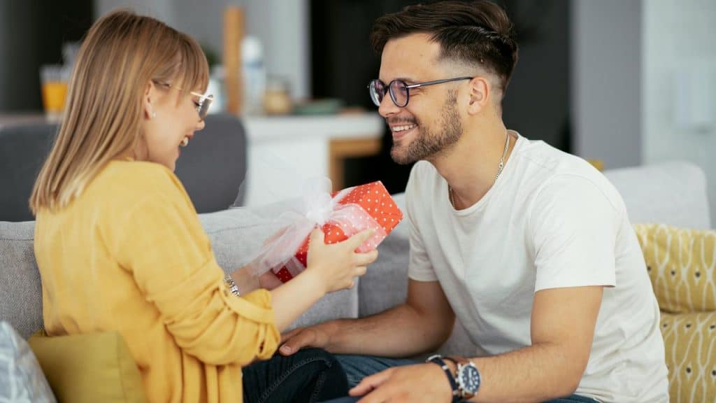 A smiling man receiving a wrapped gift from a child on a couch.