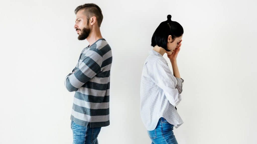 A man and woman stand back-to-back against a white wall with their arms crossed.
