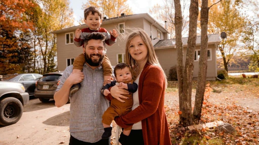 A smiling family of four stands outdoors in front of a house and autumn trees.