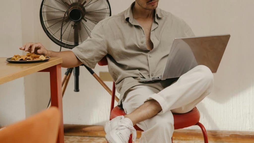 A person sitting on a chair using a laptop while reaching for snacks.