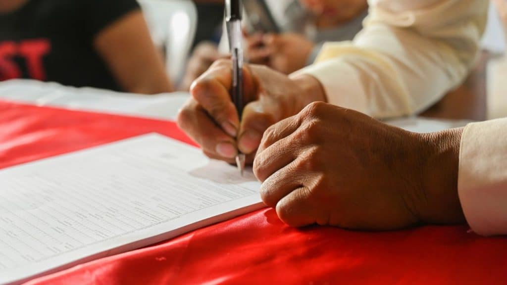 A person uses a pen to sign a document resting on a red surface cloth.