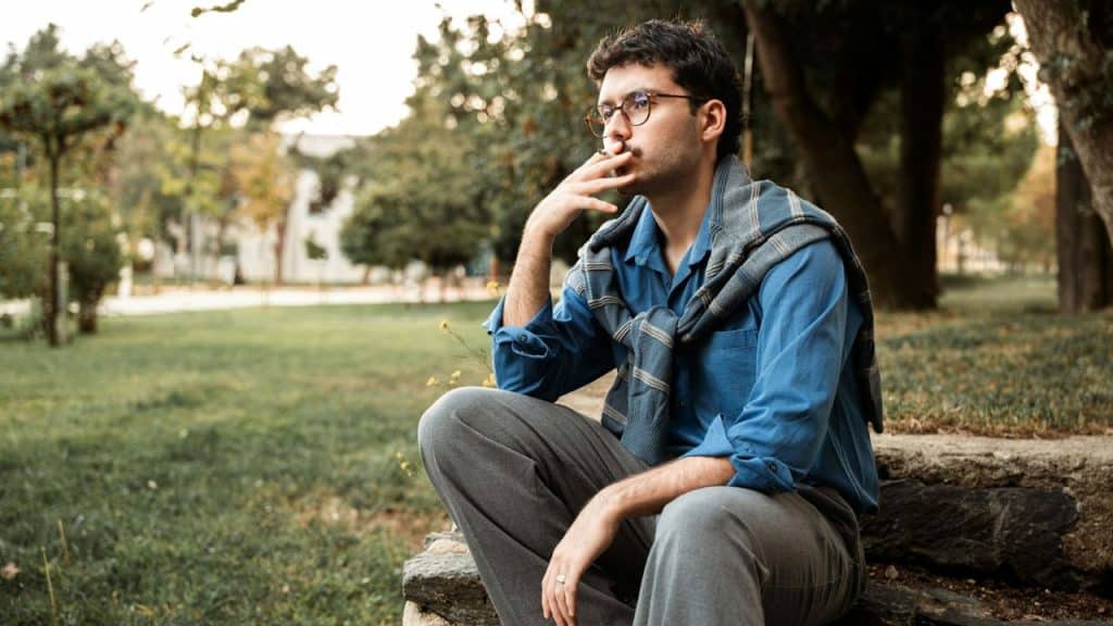 A man wearing glasses and a blue shirt sits outdoors on a stone ledge smoking.