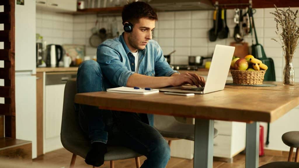 A man wearing headphones works on a laptop at a kitchen table.