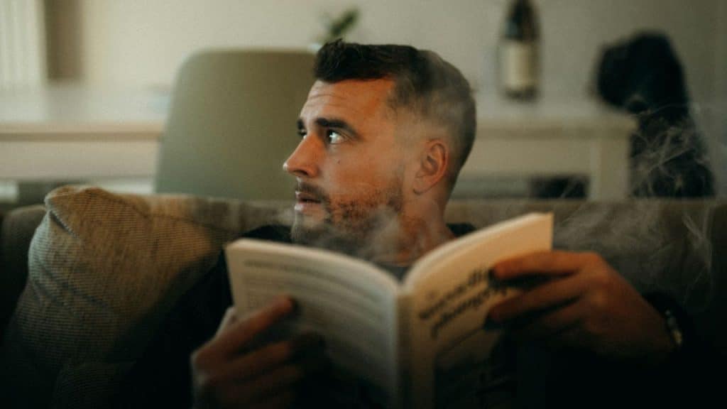 A man sits on a couch holding an open book while looking off to the side.