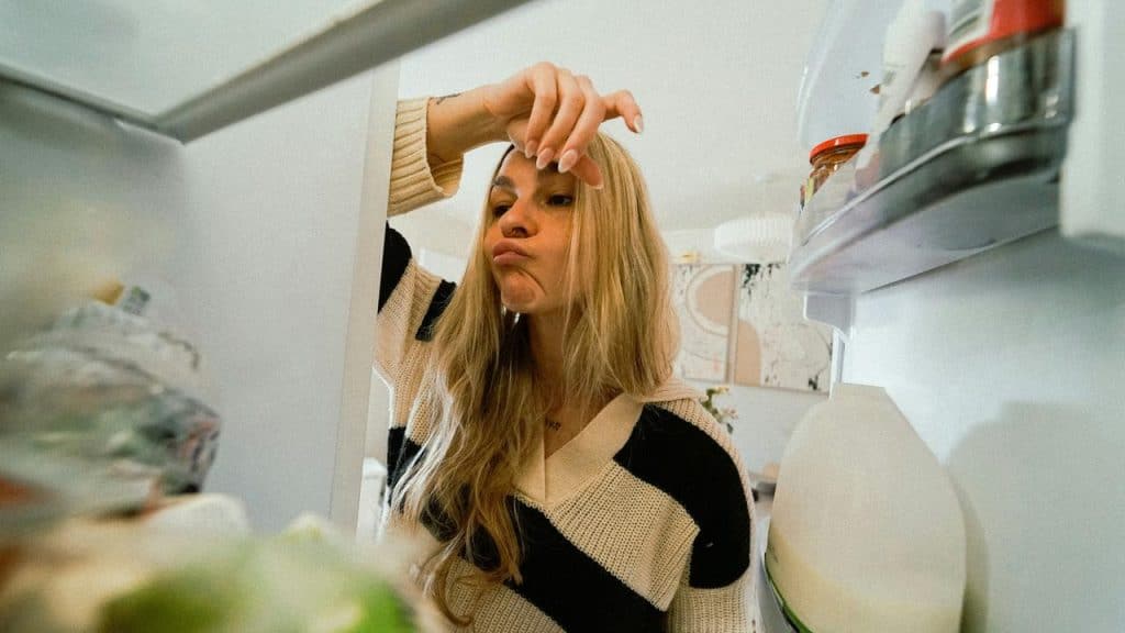 A woman peering into an open refrigerator, looking unsure.