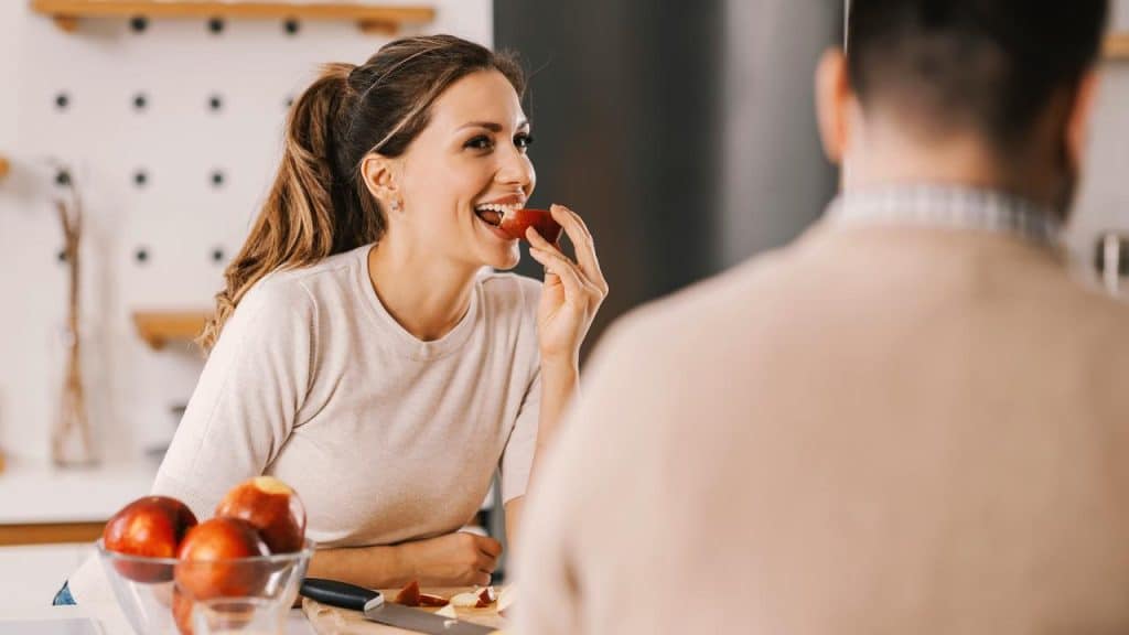 A woman smiling while eating a slice of apple in a kitchen.