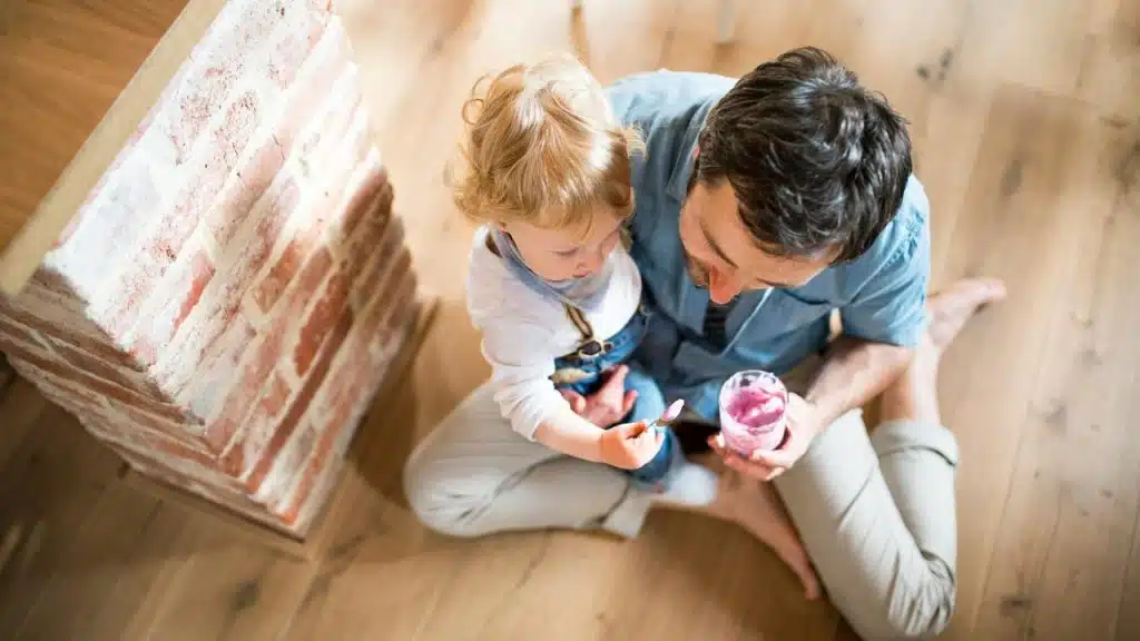 A man feeding a toddler while sitting on the floor.