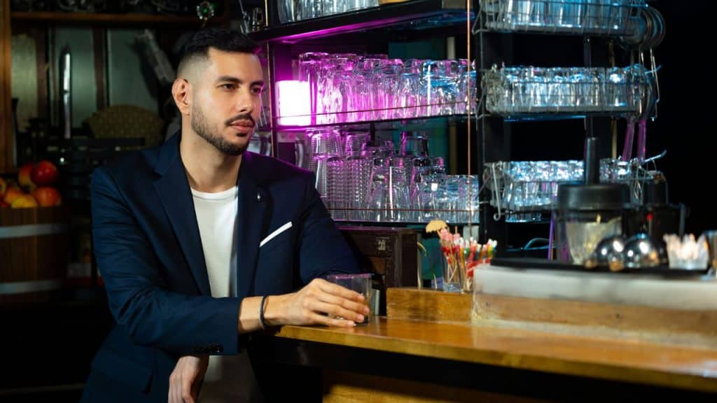 A bearded man in a navy blazer sits at a bar holding a glass of amber liquid.