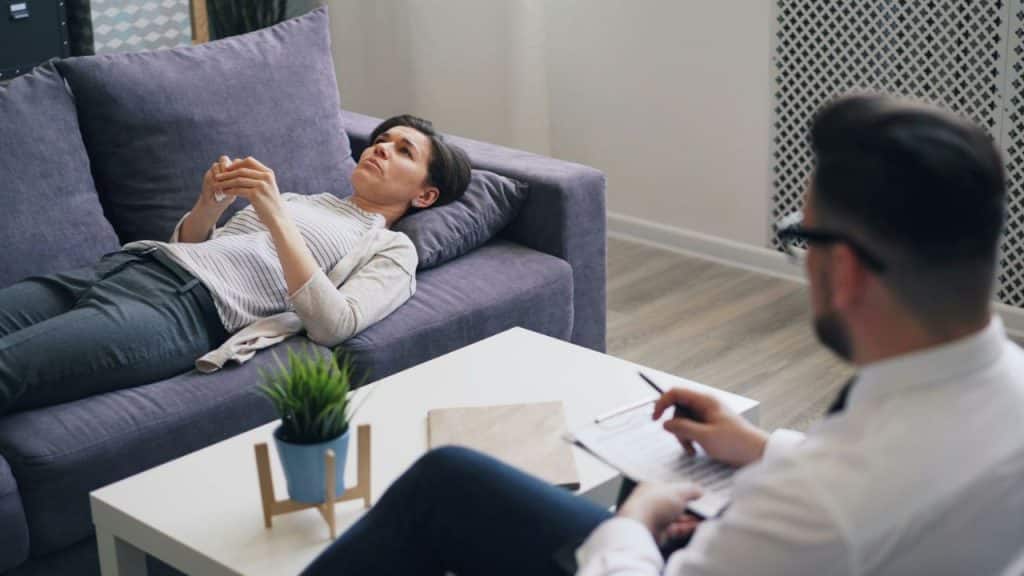 A woman lies on a purple sofa while a man takes notes on a clipboard.