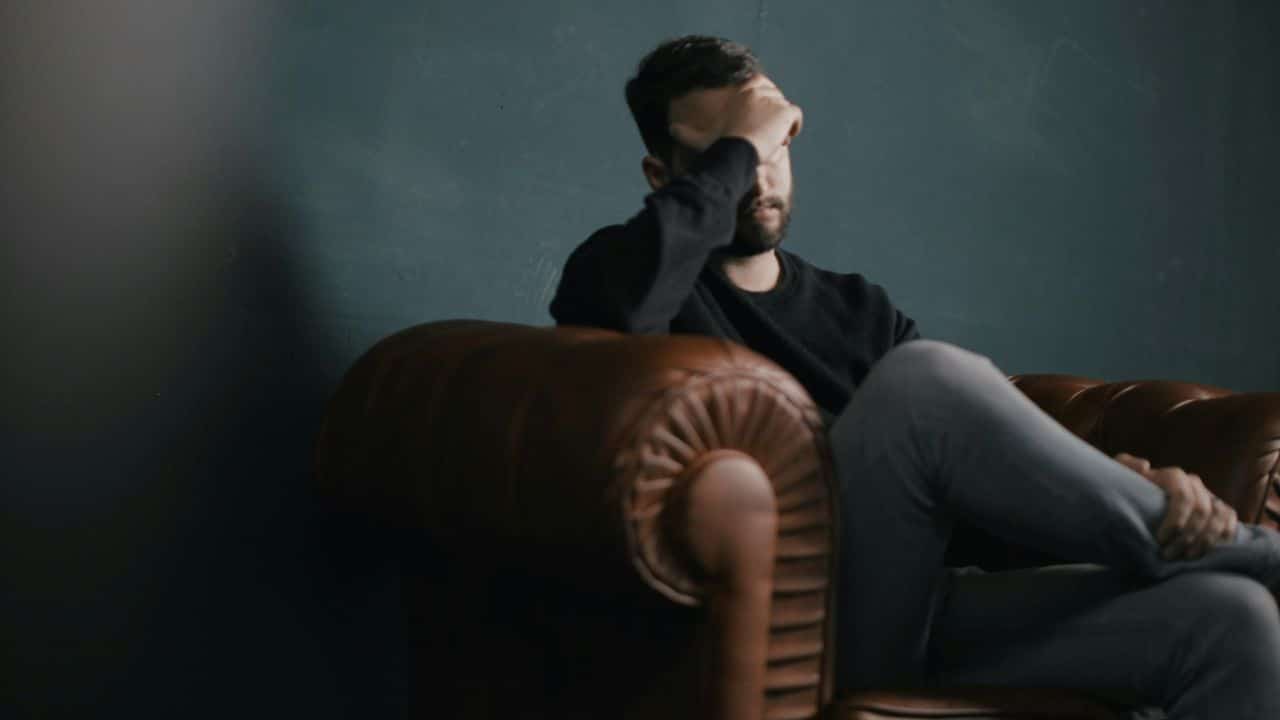 A man sits in a leather armchair with one hand covering his forehead in shadow.
