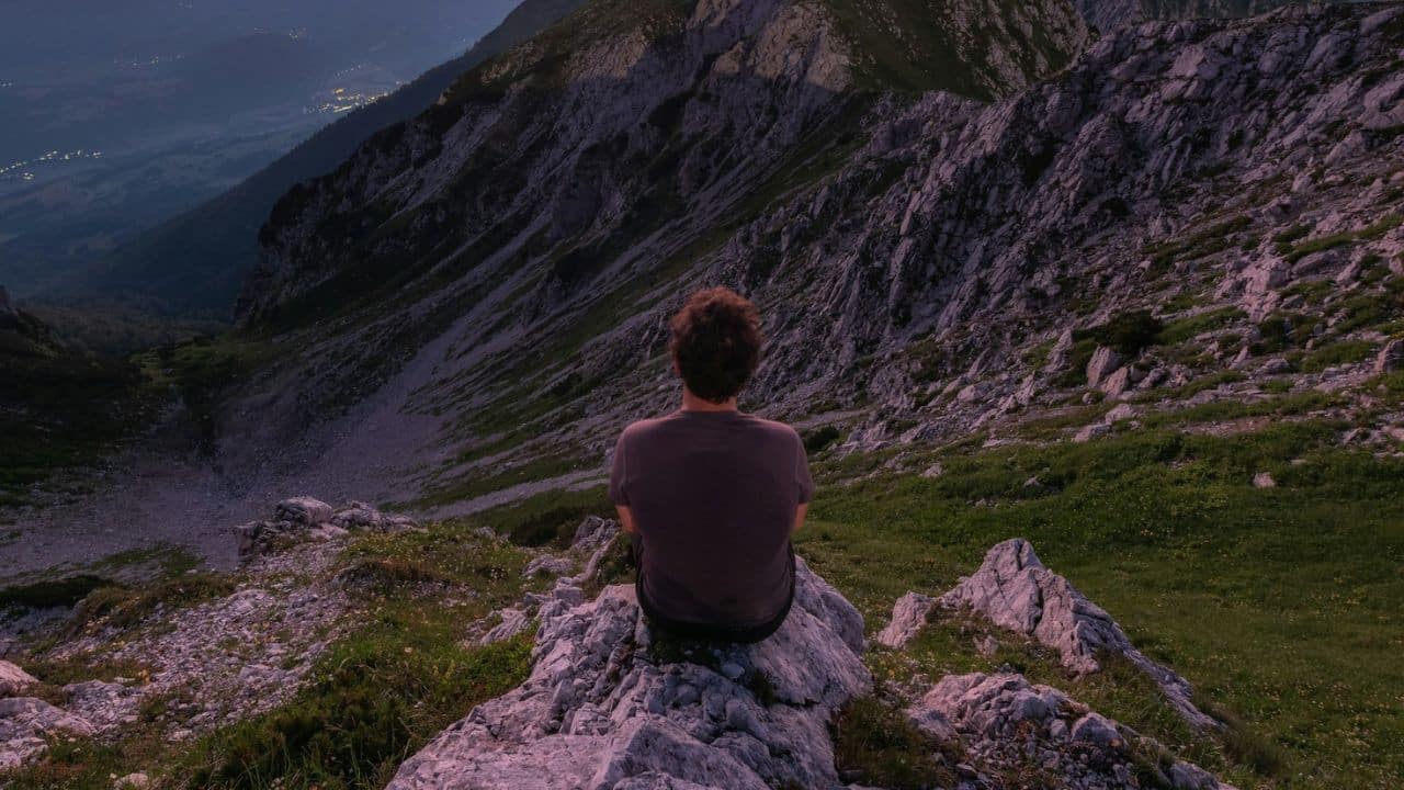 A man sits on a rocky mountain peak looking out over a valley at dusk.