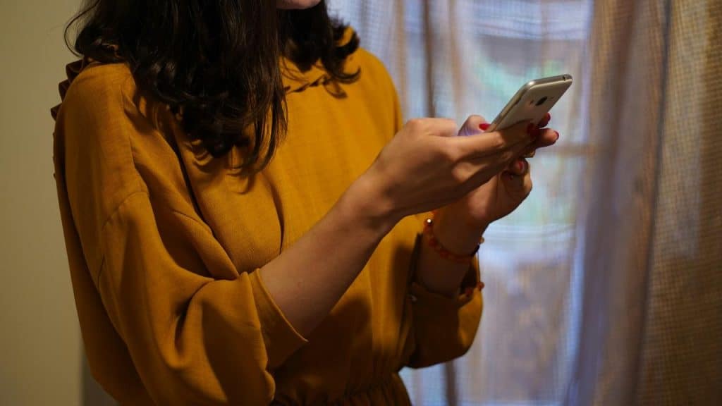 A woman in a yellow dress holding a smartphone near a curtain.
