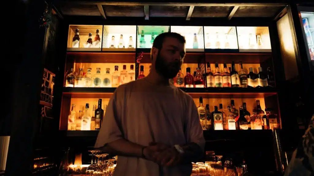 A blurred man with a beard stands behind a bar with bottles on lit shelves.