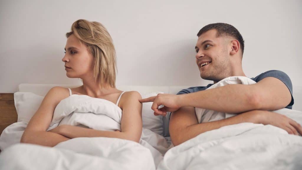 A smiling man poking a woman to get her attention as she looks away angrily while they lie in a bed together.