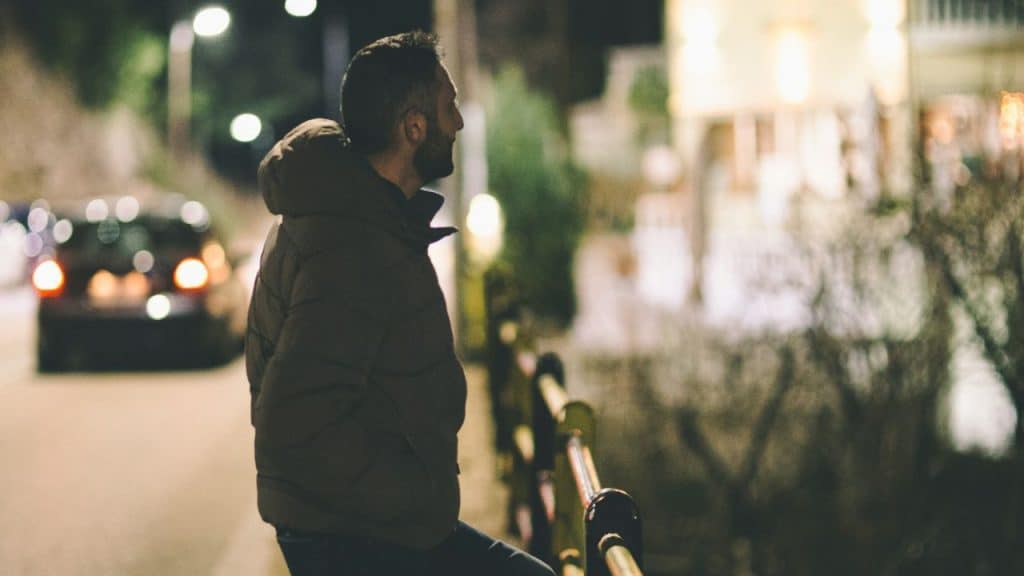 A man in a brown puffer jacket leans against a railing overlooking a street at night.