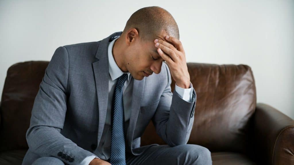 A man in a grey suit sits on a leather couch with his hand on his forehead.