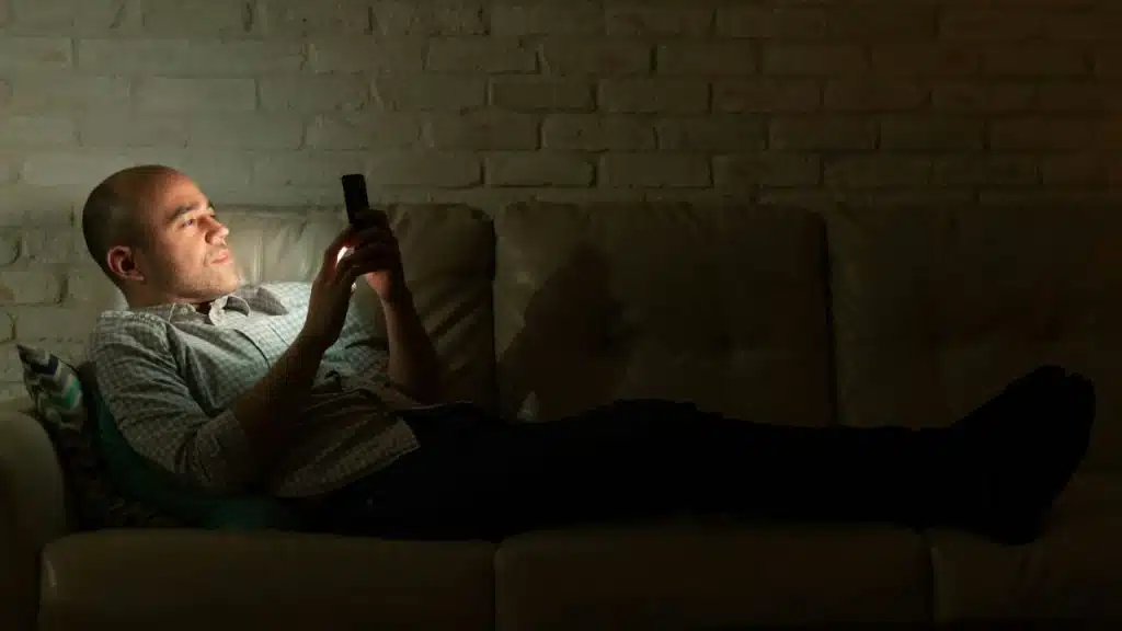 A man lies on a leather sofa in a dark room, illuminated by a smartphone.