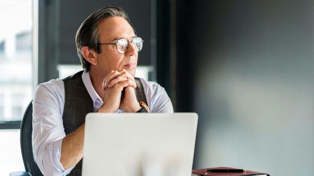 A man in glasses with his hands clasped sits behind a laptop in an office.