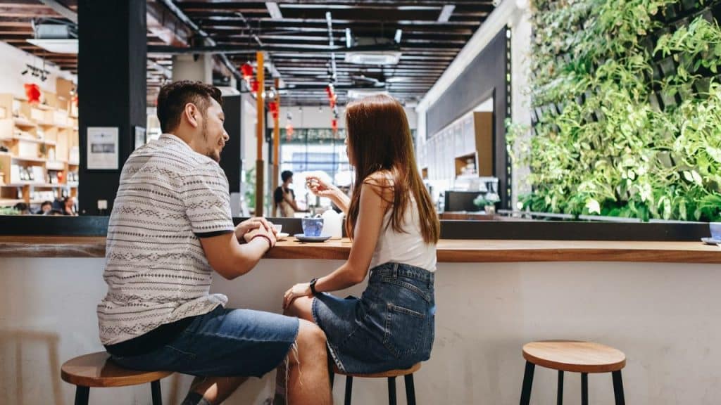 A man and woman sit at a wooden counter in a cafe, facing each other while talking.